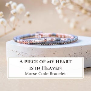 May include: A delicate bracelet featuring alternating rose gold and dark grey metallic beads arranged in a pattern. The text A PIECE OF MY HEART IS IN HEAVEN Morse Code Bracelet is displayed in a white rectangular box below the jewelry item.