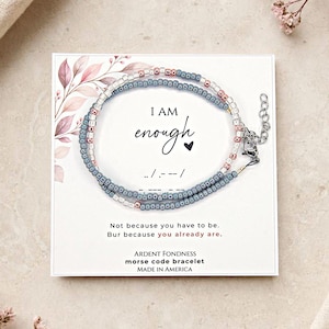 May include: A Morse code bracelet featuring small blue and metallic rose gold beads on a display card. The card reads I AM enough with a heart icon and Morse code dots and dashes. Text includes Not because you have to be. But because you already are.