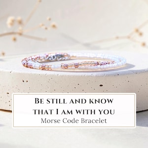 K&ouml;nnte beinhalten: Ein zartes Armband mit einem Muster aus kleinen wei&szlig;en und metallisch ros&eacute;goldenen Perlen in Morsecode-Anordnung. Ein rechteckiges Etikett unter dem Armband zeigt den Text: BE STILL AND KNOW THAT I AM WITH YOU Morse Code Bracelet.
