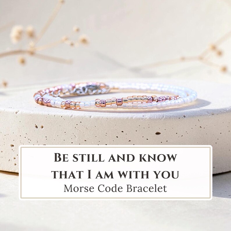 K&ouml;nnte beinhalten: Ein zartes Armband mit einem Muster aus kleinen wei&szlig;en und metallisch ros&eacute;goldenen Perlen in Morsecode-Anordnung. Ein rechteckiges Etikett unter dem Armband zeigt den Text: BE STILL AND KNOW THAT I AM WITH YOU Morse Code Bracelet.