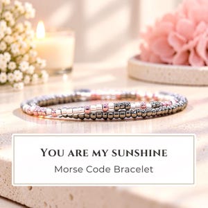 May include: A Morse code bracelet featuring a sequence of small metallic beads in shades of silver, rose gold, and dark gray. The text "YOU ARE MY SUNSHINE" and "Morse Code Bracelet" is printed in black on a white rectangular label in the foreground.