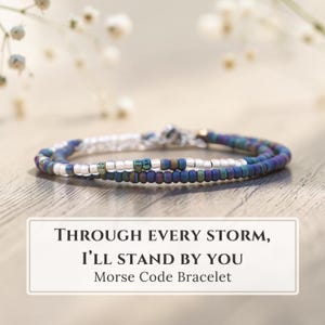 May include: A beaded bracelet featuring a pattern of matte dark blue, iridescent green, and clear glass beads. The bracelet is displayed with a card that reads: THROUGH EVERY STORM, I'LL STAND BY YOU Morse Code Bracelet. The design uses beads to represent Morse code.