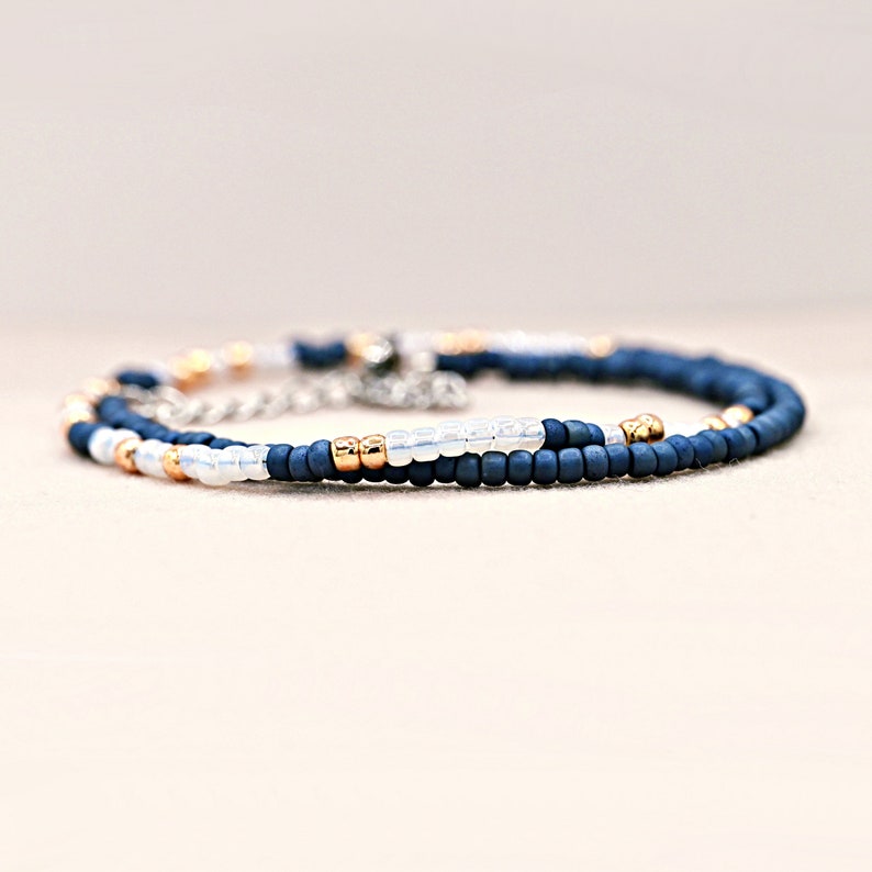 Peut inclure: Un bracelet de perles d&eacute;licat avec des perles bleu marine, dor&eacute;es et blanches. Le bracelet est compos&eacute; de petites perles rondes enfil&eacute;es ensemble, avec un fermoir en cha&icirc;ne argent&eacute;e. Le design est simple et &eacute;l&eacute;gant.