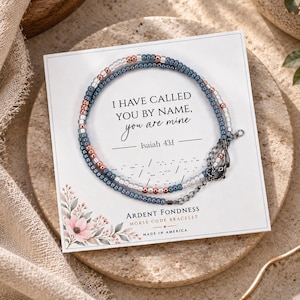 May include: A Morse code bracelet featuring blue, white, and metallic copper beads on a display card. The card reads: I HAVE CALLED YOU BY NAME, you are mine, Isaiah 43:1, Ardent Fondness Morse Code Bracelet, Made in America. The bracelet has a dark metal clasp.