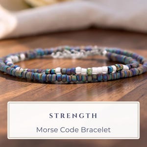 May include: A Morse code bracelet featuring iridescent blue, purple, and green seed beads interspersed with white beads. The bracelet includes a silver-toned metal clasp and is labeled with the text STRENGTH Morse Code Bracelet.