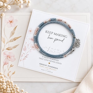 May include: A circular beaded bracelet featuring alternating blue and metallic rose gold beads, secured with a silver lobster clasp and a small flower charm. It is displayed on a white card with the text KEEP MAKING him proud and Morse code.