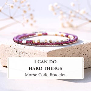 May include: A delicate beaded bracelet featuring a pattern of deep magenta, white, and metallic gold beads arranged in a sequence. The text I CAN DO HARD THINGS Morse Code Bracelet is displayed in a rectangular frame below the jewelry piece.
