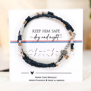 Peut inclure: Un bracelet de perles bleu marine, blanches et dor&eacute;es avec le texte "KEEP HIM SAFE day and night" et des symboles de code Morse. La carte indique "MORSE CODE BRACELET" et "MADE IN AMERICA".