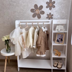 May include: A white wooden children's clothing rack with several dresses hanging on wooden hangers. The rack has shelves with a purse, picture frame, toy horse, and shoes. Decorative flower cutouts are on the wall above the rack.