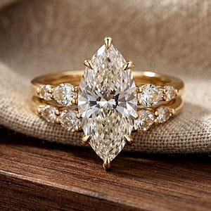 Op de afbeelding: Een gouden ringenset met een grote marquise-geslepen middensteen vastgezet met klauwen. De band is aan beide zijden versierd met kleinere peervormige stenen, wat zorgt voor een gelaagde uitstraling met een gepolijste gouden afwerking en facetten.