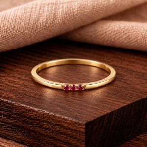 May include: A thin gold band ring featuring three small, round, vibrant red rubies set in a horizontal row. The metal has a polished, reflective finish, and the stones are held in place by tiny prongs, creating a delicate and simple jewellery piece.