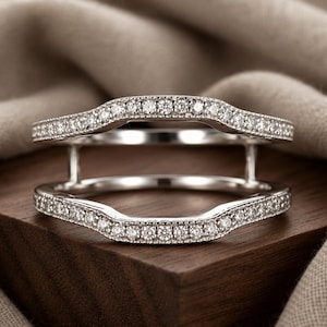 Op de afbeelding: Een zilverkleurige ringbeschermer met twee gebogen banden verbonden door verticale staven. Beide banden zijn bezet met een rij kleine, heldere, rondgeslepen stenen en afgewerkt met een delicate milgrain-rand langs de metalen randen.