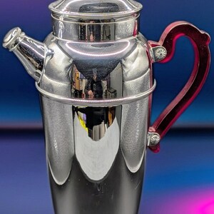 May include: A polished silver cocktail shaker with a red handle. The shaker has a spout and a lid, and it sits on a rounded base. The handle is a vibrant red color, and the overall design is sleek and classic. This barware item is perfect for mixing drinks.