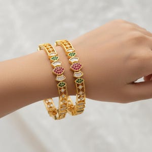 Op de afbeelding: Twee gouden armbanden met ingewikkelde ontwerpen en kleurrijke edelstenen worden getoond op de pols van een persoon. De armbanden hebben rode, groene en witte stenen, wat een vleugje elegantie aan de sieraden toevoegt.