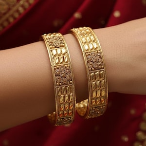 May include: Two gold bangles with detailed geometric designs are shown on a wrist. The bangles have a warm, yellow tone and a textured finish. The background is a deep red fabric, providing a contrast with the jewellery.
