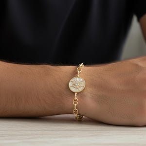 May include: Gold-toned bracelet featuring a circular pendant with a starburst design and small crystals. The bracelet has a chain link design and is worn on the wrist. The bracelet is a piece of jewelry.