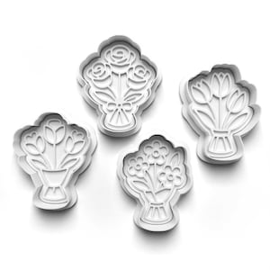 May include: Four white plastic cookie cutters in the shape of bouquets. The cutters feature designs of roses, tulips, and other flowers. Each bouquet is tied with a ribbon or has a vase-like base.