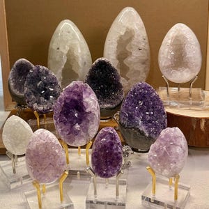 May include: A collection of amethyst and quartz crystal geodes in various sizes and shapes. The geodes are displayed on clear stands and a wooden base. The crystals range in color from deep purple to pale lavender and white.