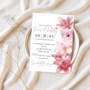 May include: A white invitation card with pink text and floral accents, announcing Ema's 16th birthday on August 21st. The card includes event details and an RSVP. The design features pink lilies and is set on a white plate and fabric.