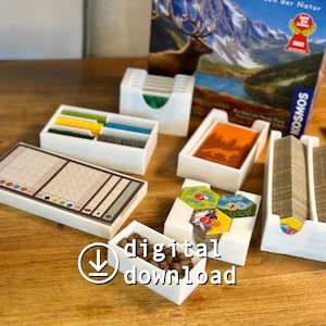 May include: A collection of white game organizers with various compartments, holding game pieces, cards, and tokens. The game pieces are in different colors. A game box with a mountain scene is in the background. The words "digital download" are in the image.
