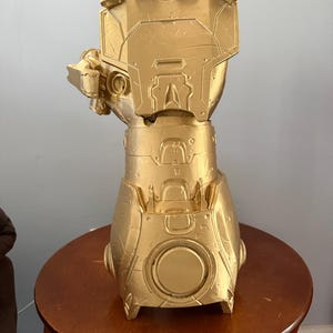 May include: A gold-coloured, detailed gauntlet sculpture. The gauntlet is made of a metallic material and has a complex, mechanical design. It is displayed on a small, round wooden table.