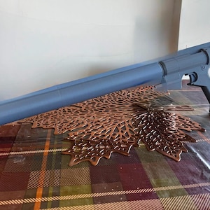 May include: A gray 3D-printed prop weapon, resembling a blaster rifle, rests on a patterned surface. The prop features a long barrel, a handle, and a black tip. A decorative copper-colored leaf-shaped placemat is beneath the prop.