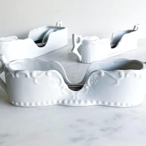 Puede incluir: Conjunto de cuatro porta cubiertos de cerámica blanca con un diseño decorativo. Los soportes tienen forma de barco y tienen un pequeño borde a cada lado para sujetar los cubiertos.