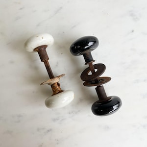 Peut inclure: Deux boutons de porte anciens, l'un blanc et l'autre noir, avec une quincaillerie métallique rouillée.