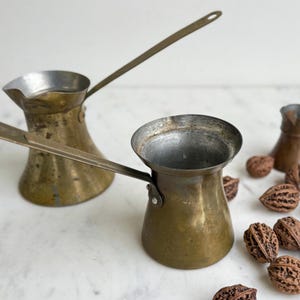 May include: Three antique brass Turkish coffee pots with long handles. The pots are on a white surface with brown nutshells scattered around them.