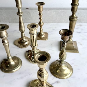 May include: A collection of antique brass candlesticks in various shapes and sizes. The candlesticks have a polished finish and are arranged on a white marble surface. The collection includes candlesticks with round, square, and hexagonal bases.