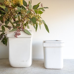 Op de afbeelding: Twee witte keramische vazen, een groter dan de andere, met een boeket groene eucalyptusbladeren en gele bloemen in de grotere vaas.