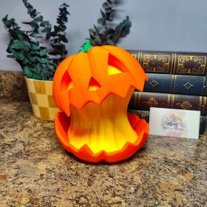 May include: An orange, 3D-printed pumpkin with a carved face, illuminated from within by a yellow light. The pumpkin is on a countertop, with a small business card and a stack of books in the background. A small plant is in a decorative pot.