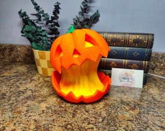 May include: An orange, 3D-printed pumpkin with a carved face, illuminated from within by a yellow light. The pumpkin is on a countertop, with a small business card and a stack of books in the background. A small plant is in a decorative pot.