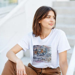 May include: White t-shirt featuring a detailed image of a tiger swimming in water. The tiger's face is visible, with orange and black stripes. The shirt is worn by a person sitting on white steps, wearing brown pants.