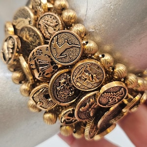 May include: A gold-toned bracelet composed of multiple circular coin-like charms featuring various embossed designs, including a rabbit, a shield, and botanical motifs, interspersed with small, textured gold-toned spherical beads throughout.