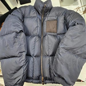 May include: A navy blue puffer jacket with a high collar and a full-length zip. The jacket features a brown square patch with a white "A" logo on the chest. The jacket has a quilted design and elasticated cuffs.