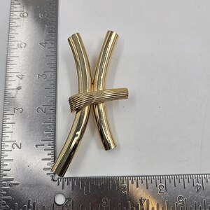 May include: A gold-tone metal brooch shaped like the Pisces zodiac symbol, featuring two curved tubes joined by a horizontal ribbed band. A metal ruler is positioned along the left and bottom edges, showing the piece is approximately 7.6 cm tall.