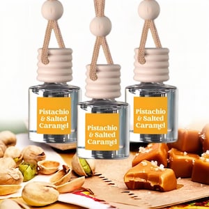 May include: Three clear glass car air fresheners with wooden tops and beige cords. Each has a yellow label with the text "Pistachio & Salted Caramel". The image also shows pistachios and caramel candies.