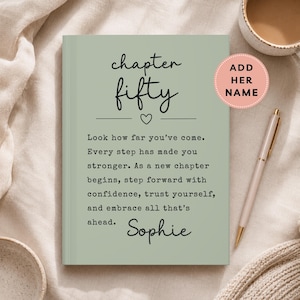 May include: Sage green journal with the words "chapter fifty" and a heart symbol. The journal includes inspirational text and the name "Sophie". A pink sticker says "Add Her Name". A pen and a mug are also in the image.