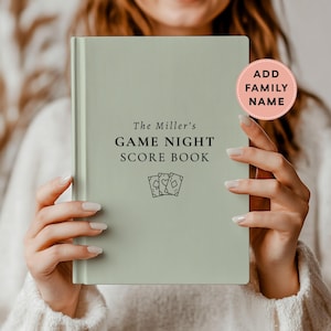 May include: A sage green hardcover score book with black text that reads The Miller's GAME NIGHT SCORE BOOK and a small illustration of three playing cards. A circular pink sticker on the top right corner says ADD FAMILY NAME.