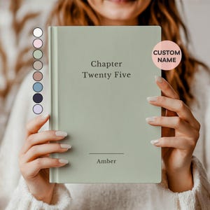 May include: A sage green hardcover journal with the text "Chapter Twenty Five" and the name "Amber" printed on the cover. A pink circle with the words "CUSTOM NAME" is on the right side. The journal is held by a person.