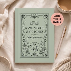 May include: A sage green hardcover book titled A LITTLE BOOK OF GAME NIGHTS & VICTORIES with The Johnsons printed below. The cover features a black decorative floral border, a hand of playing cards, and a single die illustration.