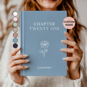 May include: A light blue book with the text "CHAPTER TWENTY ONE" and the name "ALEXANDRA" printed on the cover. A white floral illustration is below the text. A pink circle with the words "CUSTOM NAME" is on the right side of the book.