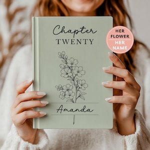 May include: A sage green hardcover book featuring the black text Chapter TWENTY and the name Amanda. A black line drawing of a floral branch is centered on the cover. A circular pink sticker in the top right corner contains the text HER FLOWER HER NAME.