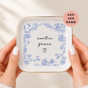 May include: A square white ceramic trinket dish with a gold-toned rim, featuring a blue floral border design. The center displays the black text "auntie grace" above a small black heart outline. A pink circular badge reads "ADD HER NAME".