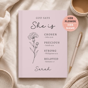 May include: A light pink hardcover book featuring a black line drawing of a carnation and the text: GOD SAYS She is CHOSEN - John 15:16, PRECIOUS - Isaiah 43:4, STRONG - Philippians 4:13, BELOVED - Romans 1:7, and the name Sarah.