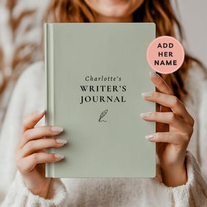 May include: A sage green hardcover journal with the text Charlotte's WRITER'S JOURNAL and a small black quill icon printed on the front cover. A circular pink badge in the upper right corner contains the black text ADD HER NAME.