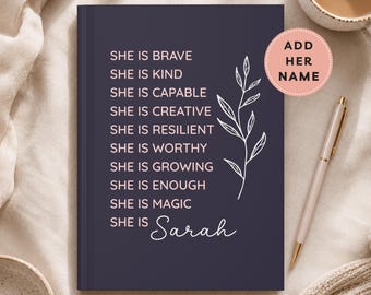 May include: A dark purple hardcover journal featuring a white line drawing of a leafy branch and light pink text that reads: SHE IS BRAVE, SHE IS KIND, SHE IS CAPABLE, SHE IS CREATIVE, SHE IS RESILIENT, SHE IS WORTHY, SHE IS GROWING, SHE IS ENOUGH, SHE IS MAGIC, SHE IS Sarah.