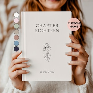May include: A white book with the text "CHAPTER EIGHTEEN" and the name "ALEXANDRA" printed in black. A black line drawing of a flower is below the text. A pink circle with the words "CUSTOM NAME" is on the right side of the book.