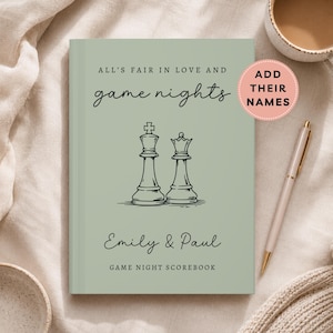 May include: A sage green scorebook with a black line drawing of a chess king and queen. The text reads: ALL'S FAIR IN LOVE AND game nights, Emily & Paul, and GAME NIGHT SCOREBOOK. A pink circular sticker says ADD THEIR NAMES.